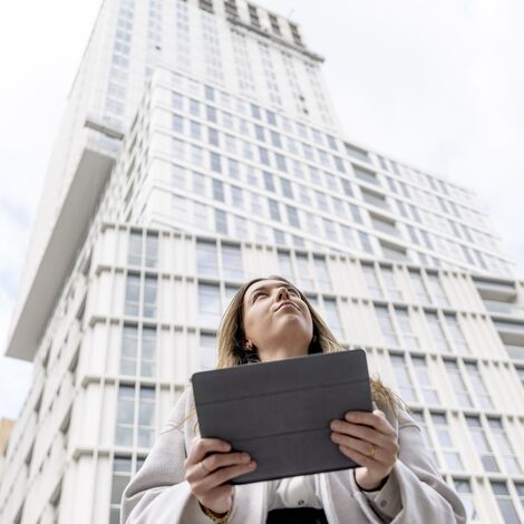 Mulher loira segurando iPad olhando para cima em direção ao prédio de apartamentos branco.