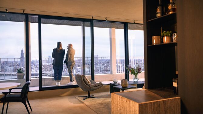 Alt: Vista interior de apartamento moderno com amplas janelas de correr e duas pessoas em uma varanda.