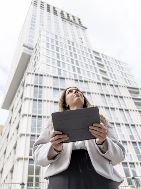 Mulher loira segurando iPad olhando para cima em direção ao prédio de apartamentos branco.
