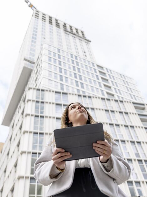 Mulher loira segurando iPad olhando para cima em direção ao prédio de apartamentos branco.